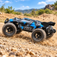 A blue and black remote control off-road truck with large knobby tires, shown driving on a rocky dirt trail with a blurred natural background of hills, trees, and blue sky.